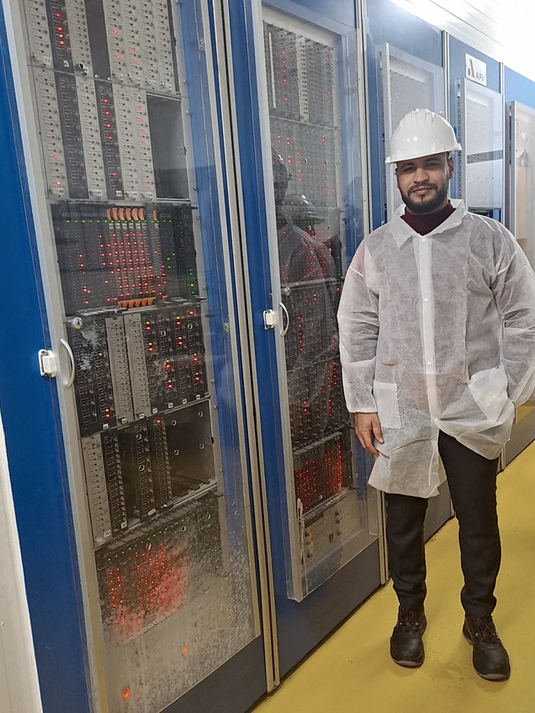 Ilyass Motya on a plant floor, beside a bank of industrial control cabinets